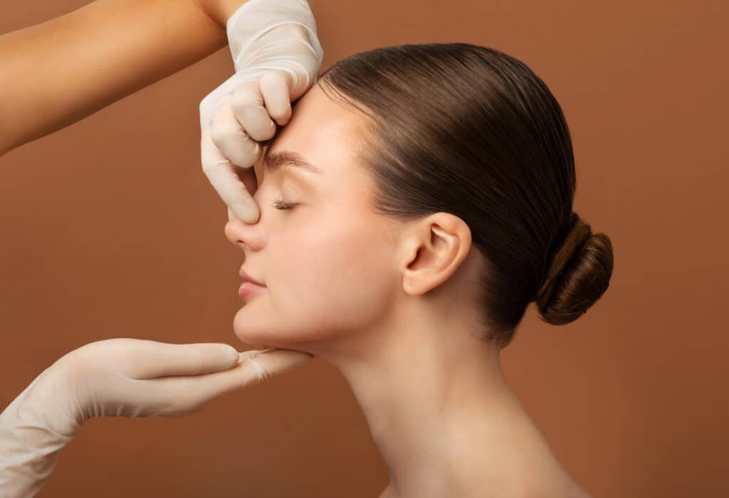Surgeon touching woman nose, consulting lady before rhinoplasty operation, brown studio background, side view. Plastic surgery concept